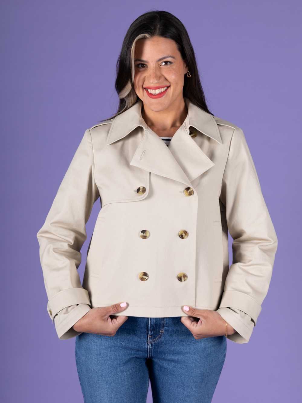 A woman modelling a light beige double-breasted, cropped trench jacket made with the Manon sewing pattern by Tilly and the Buttons. Finished with tortoiseshell buttons, paired with blue jeans, against a solid purple background.